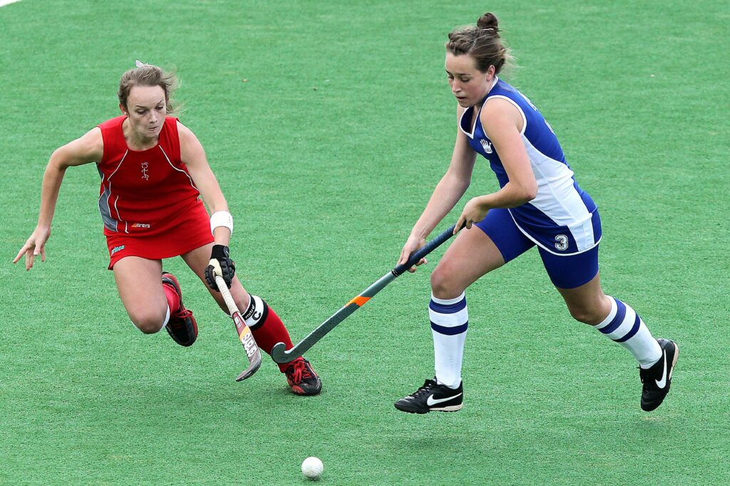 Two female athletes compete in a dynamic field hockey match on a green field.