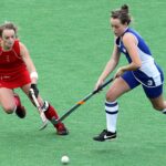 Two female athletes compete in a dynamic field hockey match on a green field.